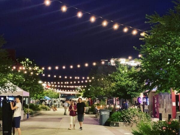 A couple strolls on a street strung with hanging lights for Bothells Wine Walk Event